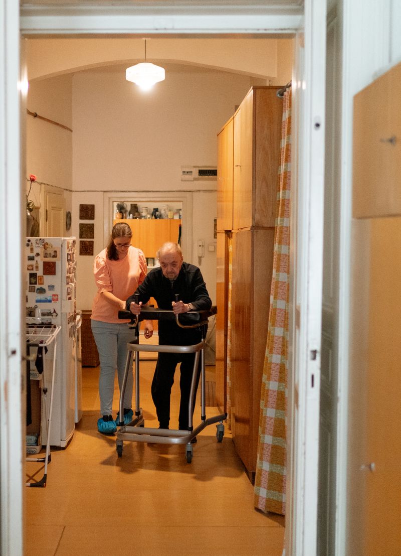 Opiekunka pomaga starszemu mężczyźnie chodzić po mieszkaniu z wysokim chodzikiem na kółkach